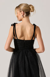 A woman is wearing a black dress with ruched fabric on the bodice, featuring thin shoulder straps that tie into bows. The skirt is made of a sheer, dotted tulle material. Her hair is styled in an elegant updo, and she is accessorized with sparkly, dangling earrings.